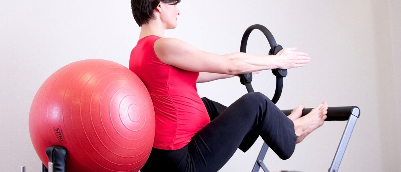 Femme enceinte assise sur le chariot d'un reformer, le dos calé contre un grand ballon, bras tendus vers un anneau de Pilates