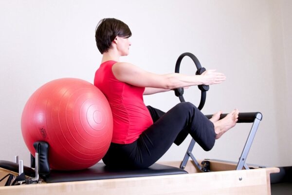 Femme enceinte assise sur le chariot d'un reformer, le dos calé contre un grand ballon, bras tendus vers un anneau de Pilates