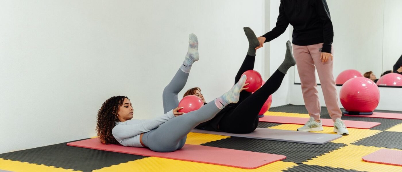 Deux femmes allongées sur des tapis, jambes tendues, avec un petit ballon de Pilates rose, guidées par une monitrice
